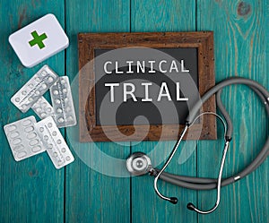 Blackboard with text & x22;Clinical trial& x22;, pills and stethoscope on blue wooden background