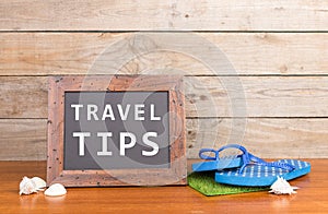 blackboard with text & x22;Travel tips& x22;, flops, seashells on brown wooden background