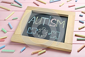 Blackboard with word Autism and drawn jigsaw puzzle pieces surrounded by colorful chalks on pink wooden background