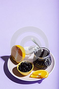 Blackcurrant jam with orange in a glass jar on a lilac background.