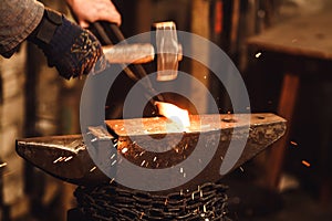 The blacksmith manually forging the red-hot metal on the anvil in smithy with spark fireworks.