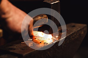 The blacksmith manually forging the red-hot metal on the anvil in smithy with spark fireworks.