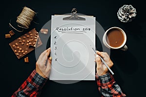 Blank album paper with New Year Resolution inscription with woman hands, pencil, chocolate, cocoa, cone on black background, top