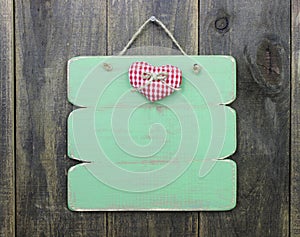 Blank antique green sign with red checkered heart hanging on rustic wood door