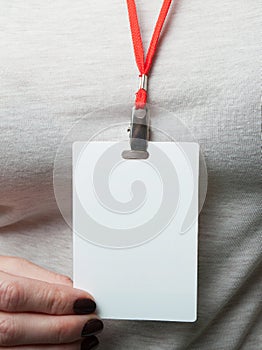 Blank badge mockup in a woman`s hand on the background of grey clothes. Plain empty name tag mock up hanging on neck with red