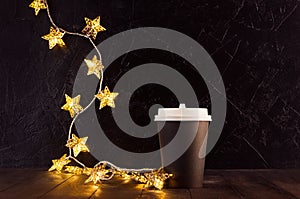 Blank black big paper coffee cup for take away with Christmas golden glowing lights with stars in dark interior, mock up.