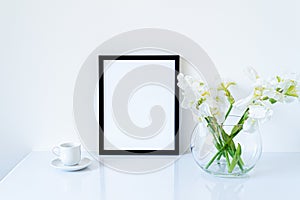 Blank black frame mockup. Fresh flowers of white irises in glass vase in the shape of a sphere on a white table and coffee cup.