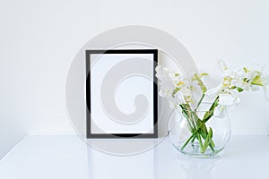 Blank black frame mockup. Fresh white irises flowers in sphere shaped glass vase on white table. White background, minimal and