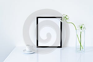 Blank black frame mockup. Small fresh flowers of white garlic allium neapolitanum in glass vase on glossy white table. White