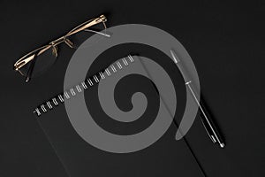 Blank black notebook, pen and glasses on dark background top view copyspace