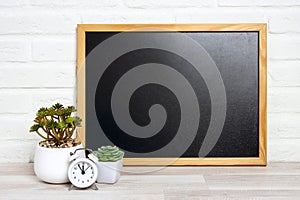 Blank  blackboard with houseplants in flowerpots and alarm clock on a table near white brick wall