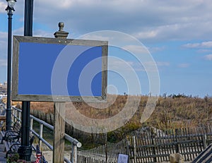 Blank blue faced sign with a wooden frame on a post on a boardwalk by brown tall grass covering a sand dune on a beach