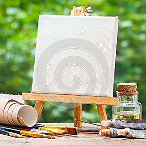 A blank canvas on easel, artistic paintbrushes and paint tubes
