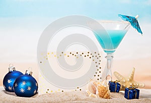 Blank card, cocktail, starfish, Christmas gifts in sand against sea.