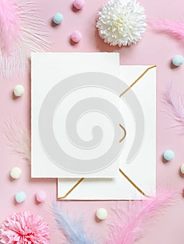 Blank card and envelope near pastel flowers, pom-poms and feathers near ring in a gift box on pink