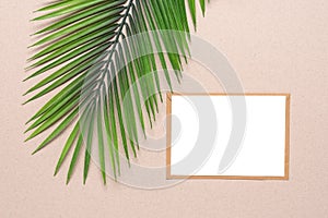 Blank card mockup, craft envelope and artificial palm leaf on beige craft paper background. Top view, flat lay