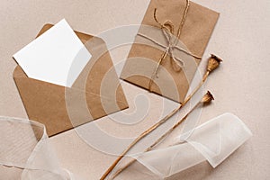 Blank card mockup in craft envelope, silk ribbon and dry poppy boxes on brown paper background. Feminine wedding invitation