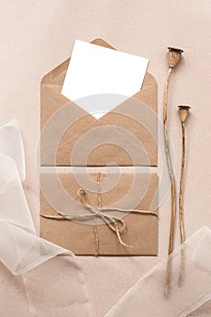 Blank card mockup in craft envelope, silk ribbon and dry poppy boxes on brown paper background. Feminine wedding invitation
