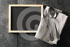 Blank, empty, black chalkboard with spoon and fork silverware and gray dish towel flat lay from above on black wooden table