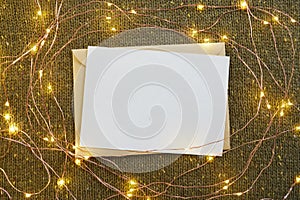 Blank greeting card, craft envelope and Christmas glowing lights on green knitted background