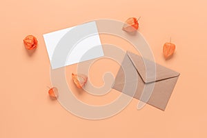 Blank greeting card mockup with brown envelope and physalis winter cherry. Pink peachy pastel background. Wedding template. Top