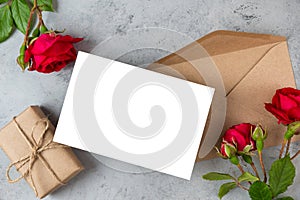 Blank greeting card with red rose flowers and gift box on gray background. Wedding invitation. Mock up. Flat lay. Womans