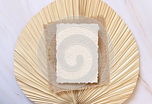 Blank handmade paper card on dry beige palm leaf near hare`s tail grass top view,  boho wedding mockup