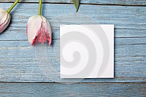 Blank invitation card mockup, white square greeting card with pink flower on blue wooden background, flat lay. Empty invitation
