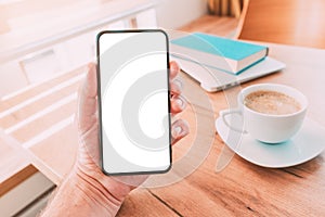 Blank mobile smart phone screen mock up. Man holding smartphone over desk with coffee cup at home