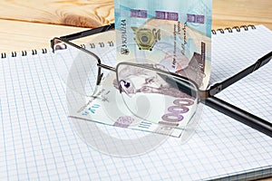 Blank notebook, glasses and ukrainian money on a wooden background. Banknote of 1000 hryvnia. Hryvnia UAH.. Close-up