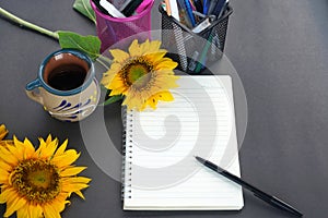 Blank notebook page with pen, sunflowers, cup of coffee and pens container on the gray table background. Business work from home.