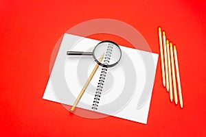 Blank notebook and pencil on red background. Magnifying glass on top of notepad