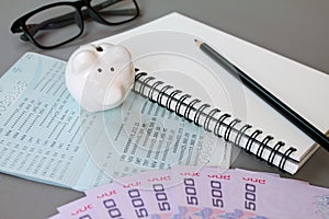 Blank notebook, pencil, savings account passbook, eye glasses, Thai money and piggy bank on gray background