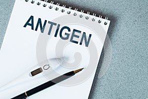 Blank notebook with thermometre and pen on table. ANTIGEN text