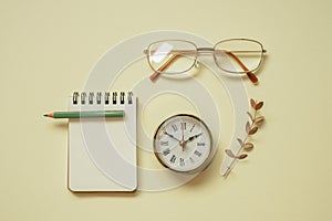 Blank notepad, pencil, clock, eyeglasses on yellow desk background. flat lay, top view, copy space