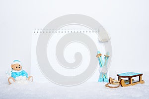 Blank notepad, teddy bear with snowballs, toy wooden blue skis, sledges and white hat on snowy background