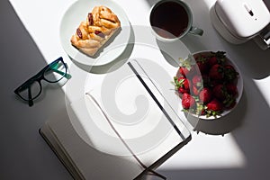 Blank open notepad with glasses, cup of tea, tea pot and strawberries in home office on white background