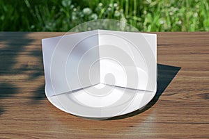 Blank opened square bifold card on a classic empty white plate on the  outdoor wooden table and green grass behind, clean template