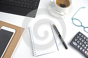 The blank page notebook on white desktop with pen, coffee, laptop, book, calculator, glasses and mobile phone