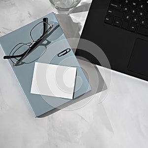 Blank paper business card mockup, light blue notebook, eyeglasses, black laptop keyboard on gray marble neutral desktop