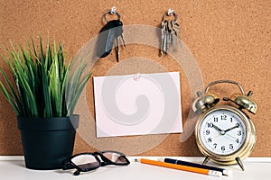 Blank paper note on cork board with car key, golden alarm clock, reading glasses, pen and green plant in pot