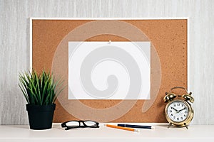 Blank paper note on cork board with golden alarm clock, reading glasses, pen and green plant in pot