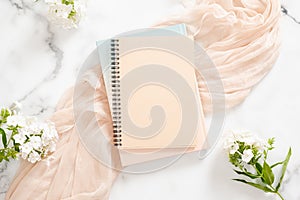 Blank paper notebook, white flowers, pastel blanket on marble background. Flat lay, top view home office desk. Beauty blogger