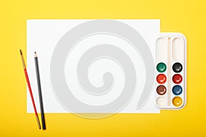 Blank paper with pencil, brush and paints on yellow background. Artist workplace concept with copy space for your ideas.