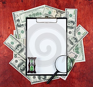 Blank paper sheet in clipboard placed on center of money dollar and wood background, sandglass and magnifier, business concept and