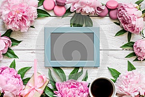 Blank photo card in frame made of pink peony flowers, macarons and coffee cup on white wooden background. flat lay