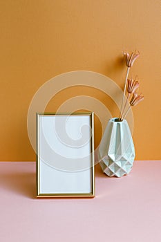 Blank photo frame with vase of dry flowers on pink table. orange wall background