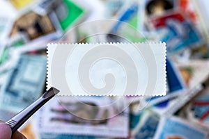 Blank postage stamp in tweezers against blurred background collection of multicolored postage stamps of different countries