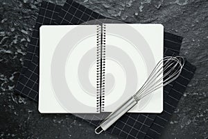 Blank recipe book and whisk on black textured table, flat lay. Space for text