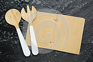 Blank recipe book and wooden utensils on black textured table, flat lay. Space for text